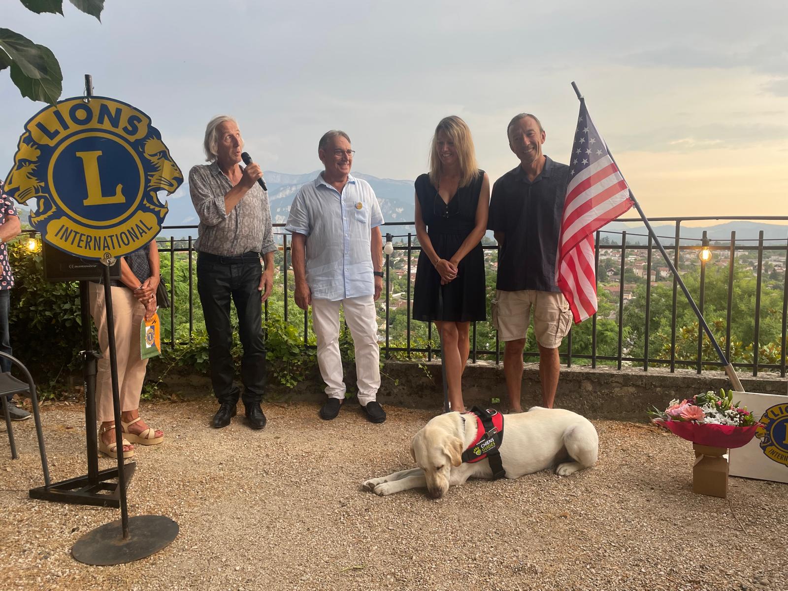 Magali Escoffier prend la parole avec son chien-guide Vax couché à ses pieds, entourée des membres du Lions Club de Voiron